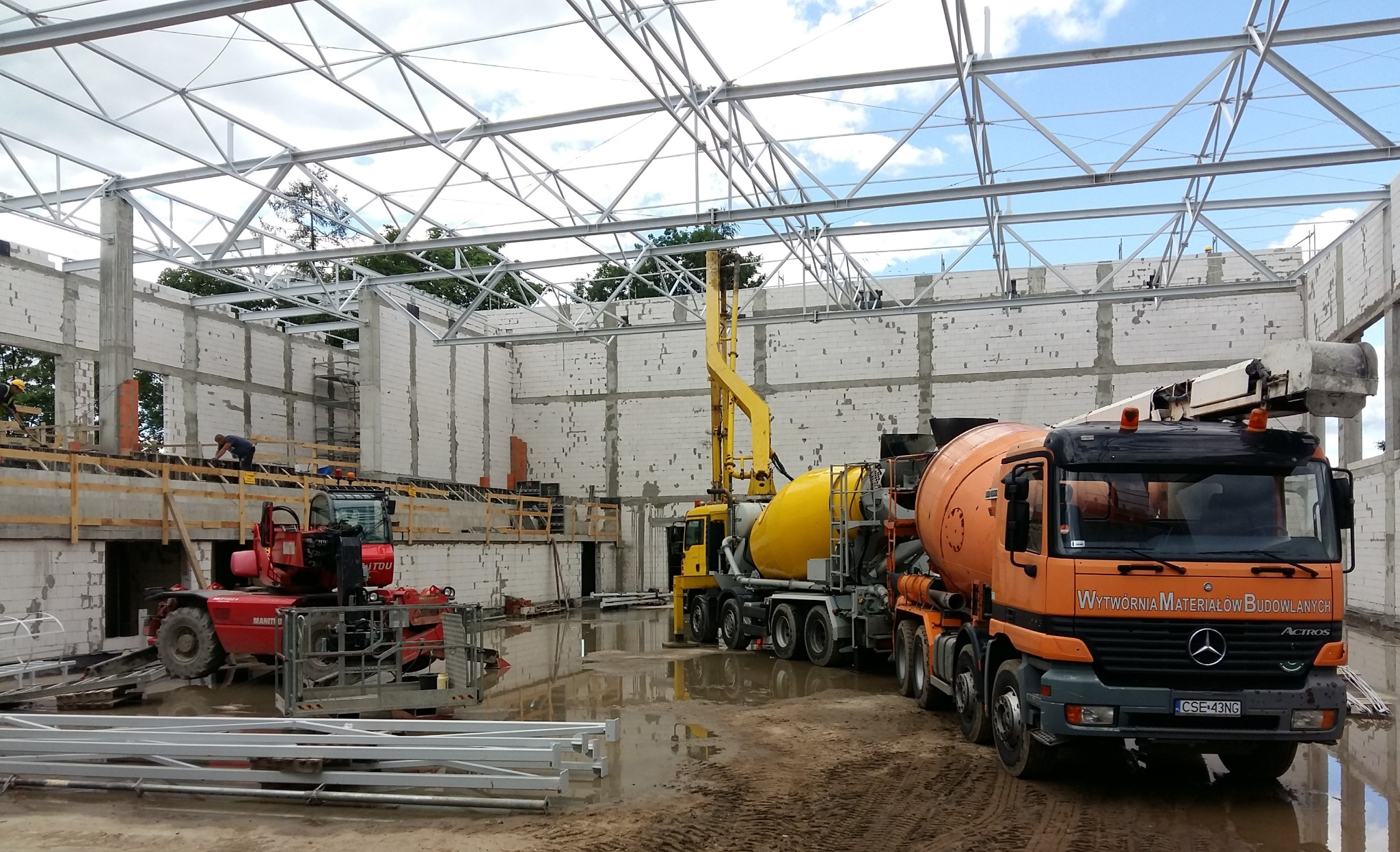 Budowa hali: Pomarańczowa betonomieszarka marki Mercedes, żółta pompa do betonu i czerwony wózek widłowy na mokrym placu budowy z widoczną konstrukcją stalową dachu i ścianami z bloczków betonowych.