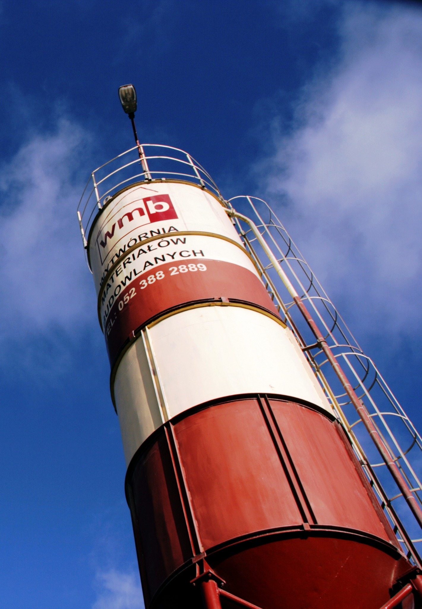 Silos betoniarni WMB w Sępólnie Krajeńskim, z widocznym logo i numerem telefonu, sfotografowany pod kątem w słoneczny dzień na tle błękitnego nieba.