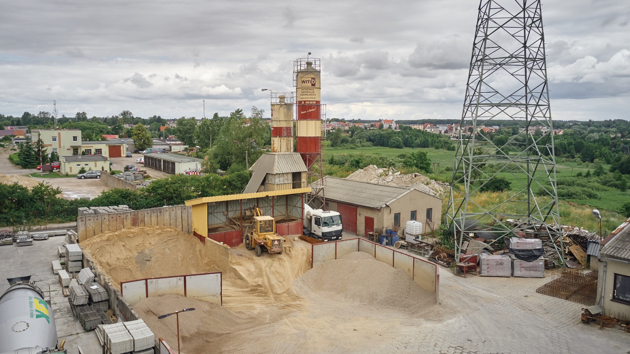 Widok z góry na teren betoniarni z silosami, stertami piasku i żwiru, ładowarką, betonomieszarką oraz elementami infrastruktury energetycznej w tle.