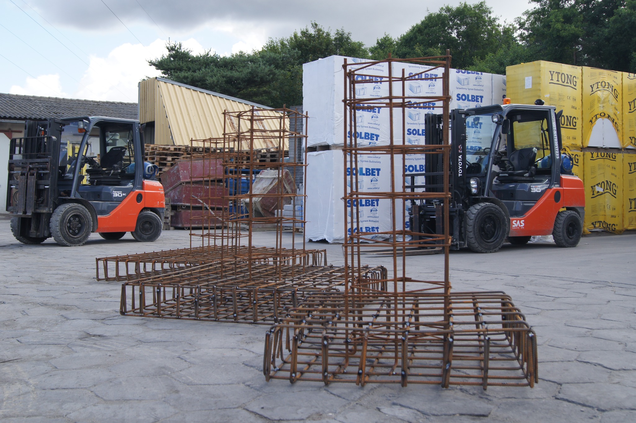 Szkielety zbrojeniowe z drutu zbrojeniowego na placu budowy, w tle wózki widłowe i palety z materiałami budowlanymi SOLBET i YTONG.