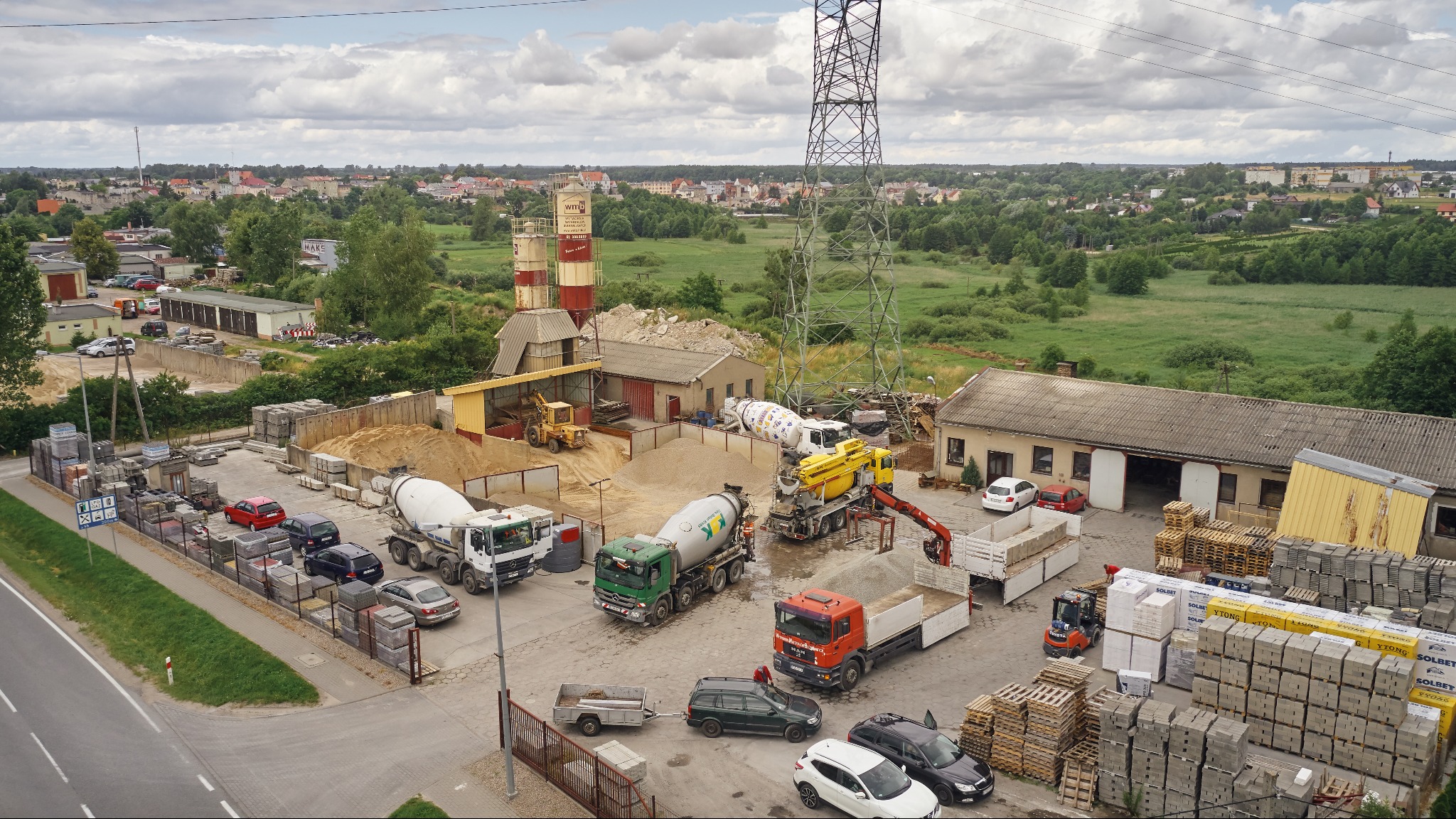 Widok z góry na plac betoniarni z zaparkowanymi samochodami, betoniarkami, koparką i stertami materiałów budowlanych na paletach, w tle zabudowa miejska i słup wysokiego napięcia.