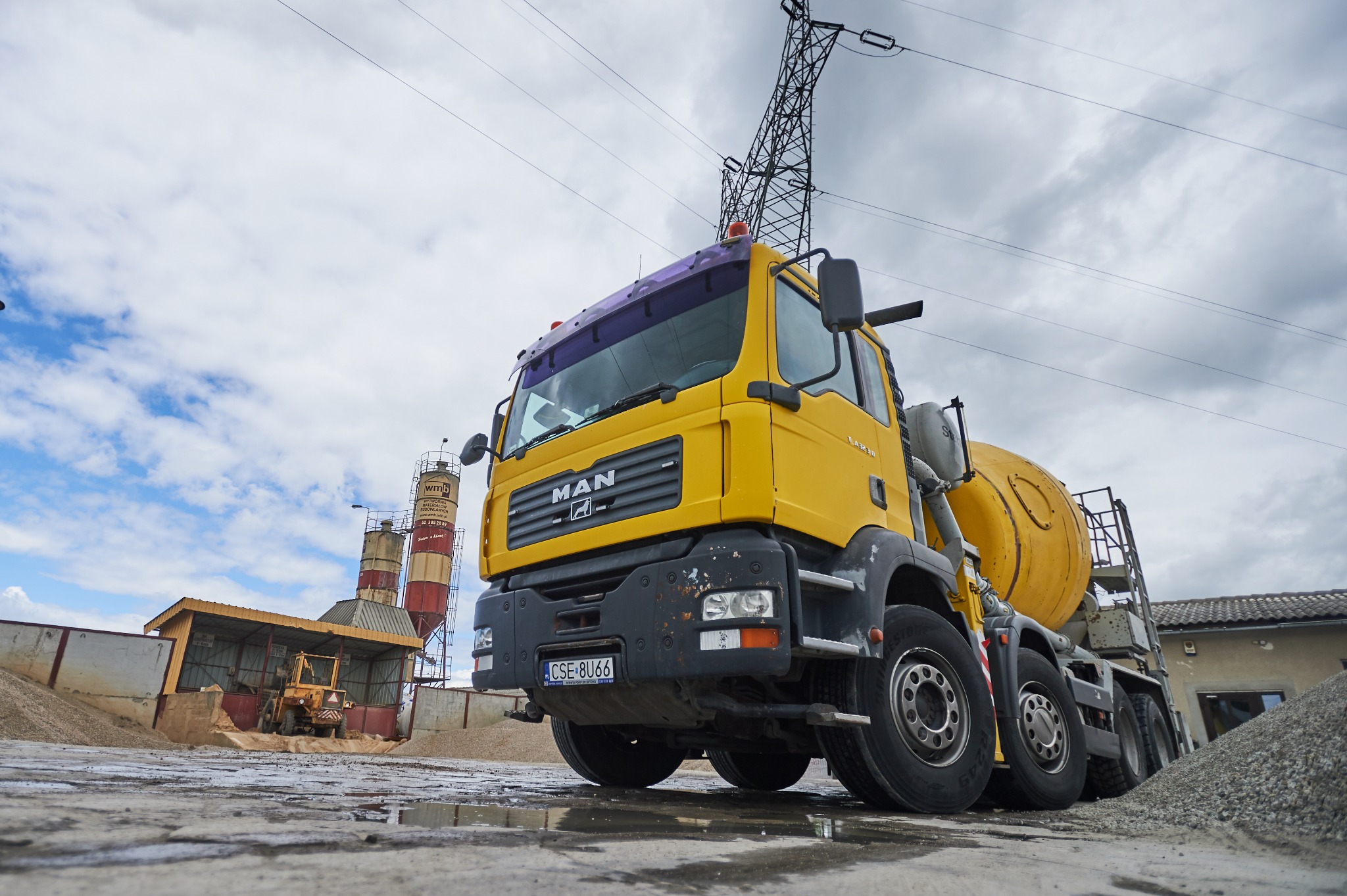 Żółta betonomieszarka MAN na terenie zakładu, w tle silosy i hałda żwiru, widoczne ślady wilgoci na betonowej nawierzchni.