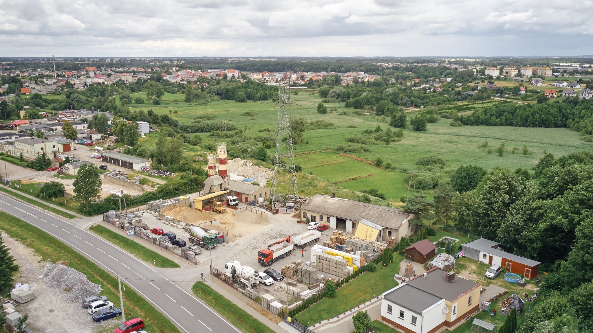 Widok z lotu ptaka na teren betoniarni z zapleczem, składowiskiem materiałów budowlanych, betonowozami, piaskiem i żwirem na tle zabudowy miejskiej i pól.