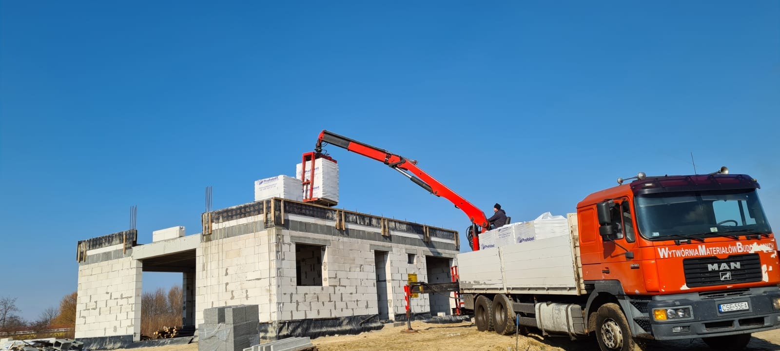 Czerwony dźwig samochodowy umieszcza pakiet białych bloczków na budowie domu z szarej kostki. W tle błękitne niebo.