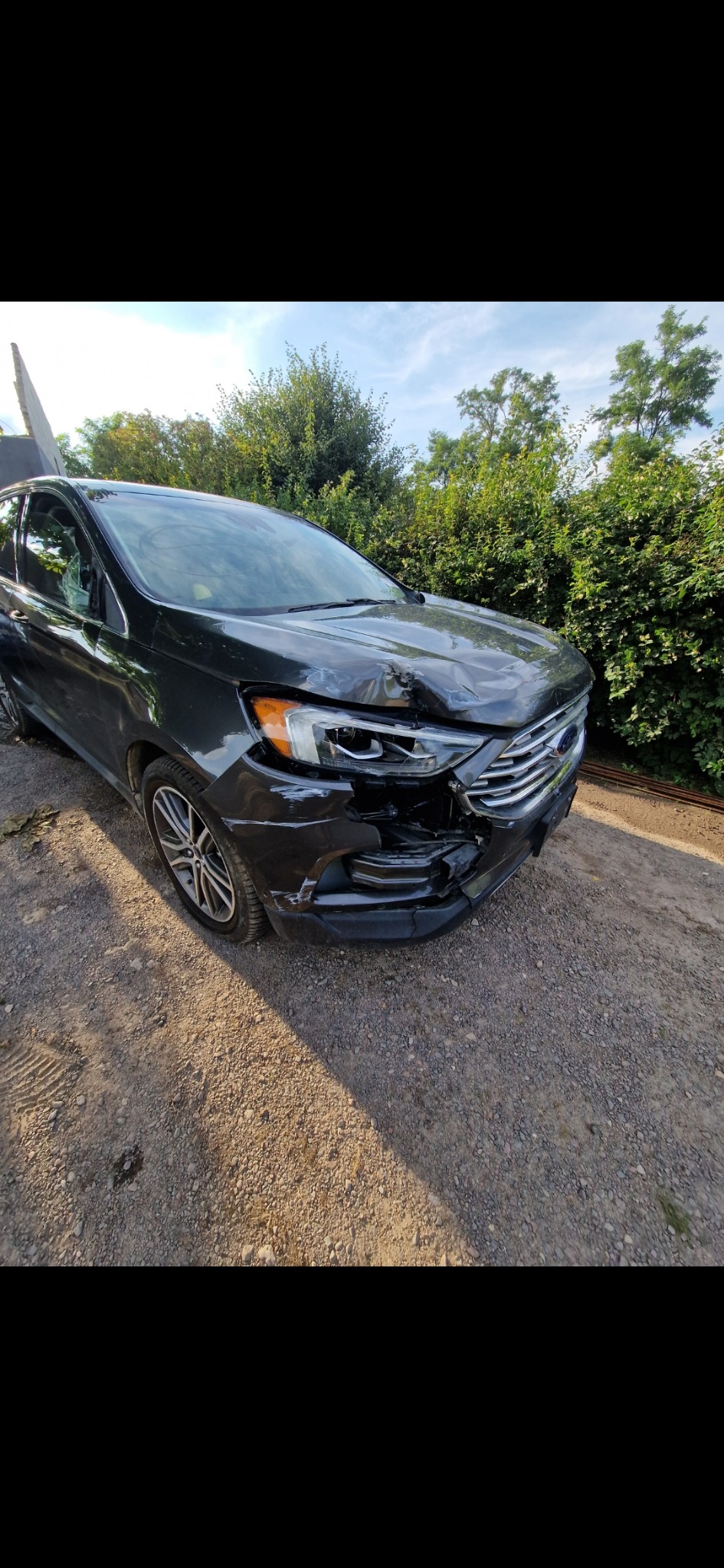 Uszkodzony przód czarnego samochodu Ford Edge na żwirowym podłożu, widoczne wgniecenia na masce i zniszczony zderzak, w tle zieleń.