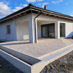 TIK BUD IVAN KIT - Nowoczesny taras z kostki brukowej w Bielsku-Białej, z widoczną elewacją domu i pracującym mężczyzną w tle, wykonany w słoneczny dzień.