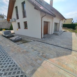 TIK BUD IVAN KIT - Nowo ułożona, wielobarwna kostka brukowa przed domem jednorodzinnym z widocznym systemem odprowadzania wody i fragmentem ażurowej nawierzchni.