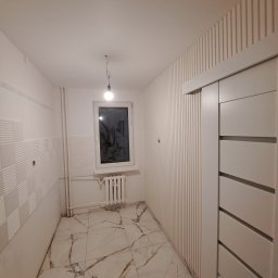 TIK BUD IVAN KIT - Wnętrze pokoju po remoncie: białe ściany z dekoracyjnymi panelami pionowymi i kwadratowymi, marmurowa podłoga, białe drzwi przesuwne i okno z grzejnikiem.