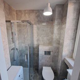 TIK BUD IVAN KIT - Wnętrze nowoczesnej łazienki z kabiną prysznicową, pralką, umywalką i podwieszaną toaletą, wykończonej szarymi płytkami imitującymi marmur, oświetlonej pojedynczą lampą wiszącą.