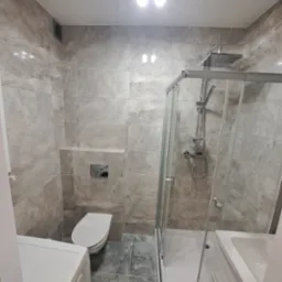 Nowoczesna łazienka po remoncie z kabiną prysznicową, podwieszaną toaletą i płytkami imitującymi marmur, oświetlona górnym światłem.