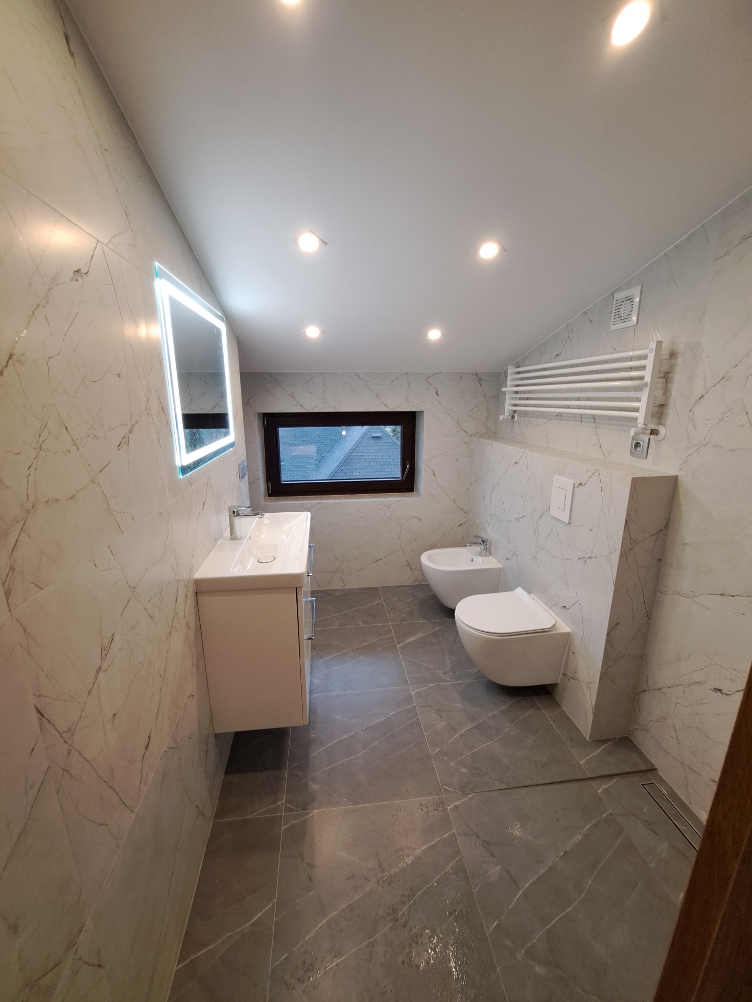Nowoczesna łazienka z marmurowymi płytkami, podwieszaną toaletą i bidetem, oświetlonym lustrem oraz grzejnikiem drabinkowym.