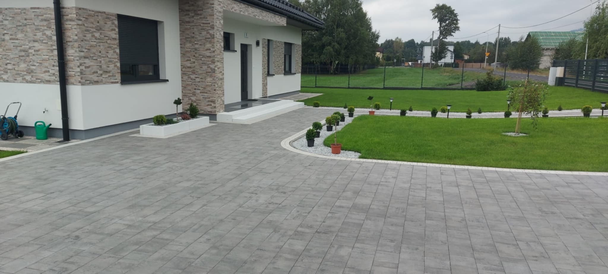Ułożona szara kostka brukowa prowadząca do nowoczesnego domu z białymi ścianami i elementami z kamienia, z zadbanym trawnikiem i rabatami z roślinami.