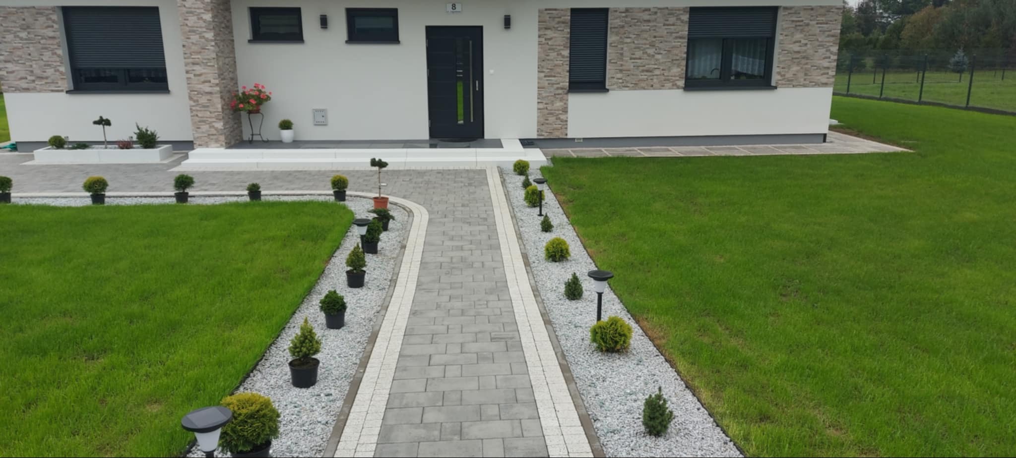 Nowoczesny dom jednorodzinny z szarą kostką brukową na ścieżce prowadzącej do drzwi, obrzeża z jasnych kostek, trawnik z równo przystrzyżoną trawą i ozdobne krzewy w doniczkach wzdłuż ścieżki.