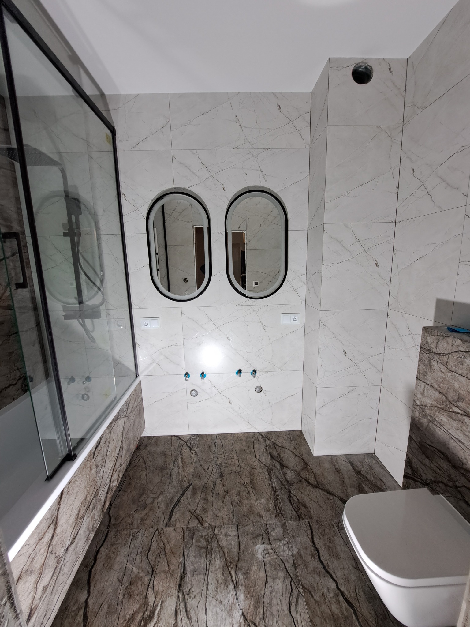 Wykończona łazienka z marmurowymi płytkami na ścianach i podłodze, owalnymi lustrami z podświetleniem, kabiną prysznicową z czarną ramą i podwieszaną toaletą.