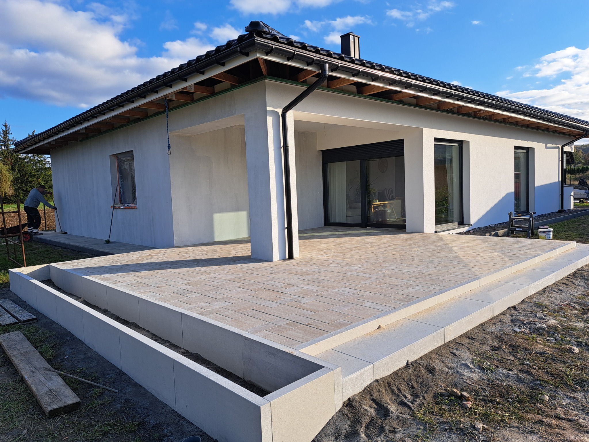 Nowoczesny taras z kostki brukowej w Bielsku-Białej, z widoczną elewacją domu i pracującym mężczyzną w tle, wykonany w słoneczny dzień.