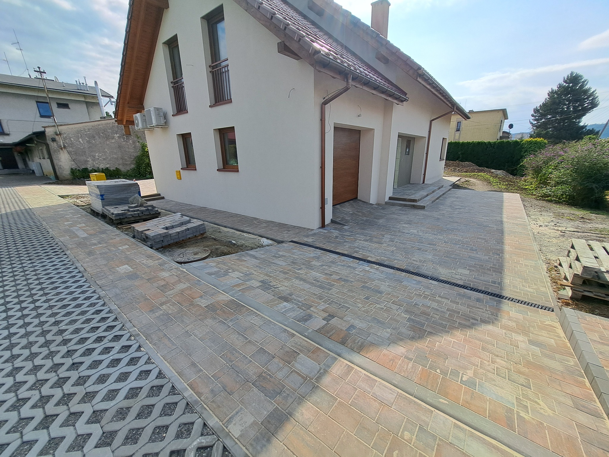 Nowo ułożona, wielobarwna kostka brukowa przed domem jednorodzinnym z widocznym systemem odprowadzania wody i fragmentem ażurowej nawierzchni.