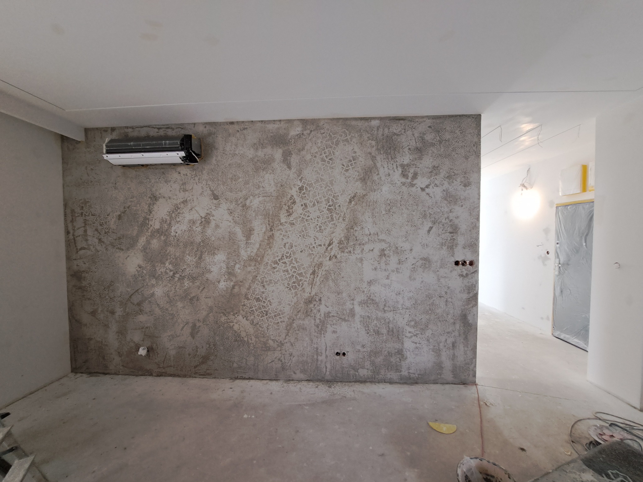 Pokój w trakcie prac wykończeniowych: ściana z dekoracyjną fakturą, zamontowany klimatyzator, widoczne puszki elektryczne i elementy instalacji w sąsiednim pomieszczeniu.