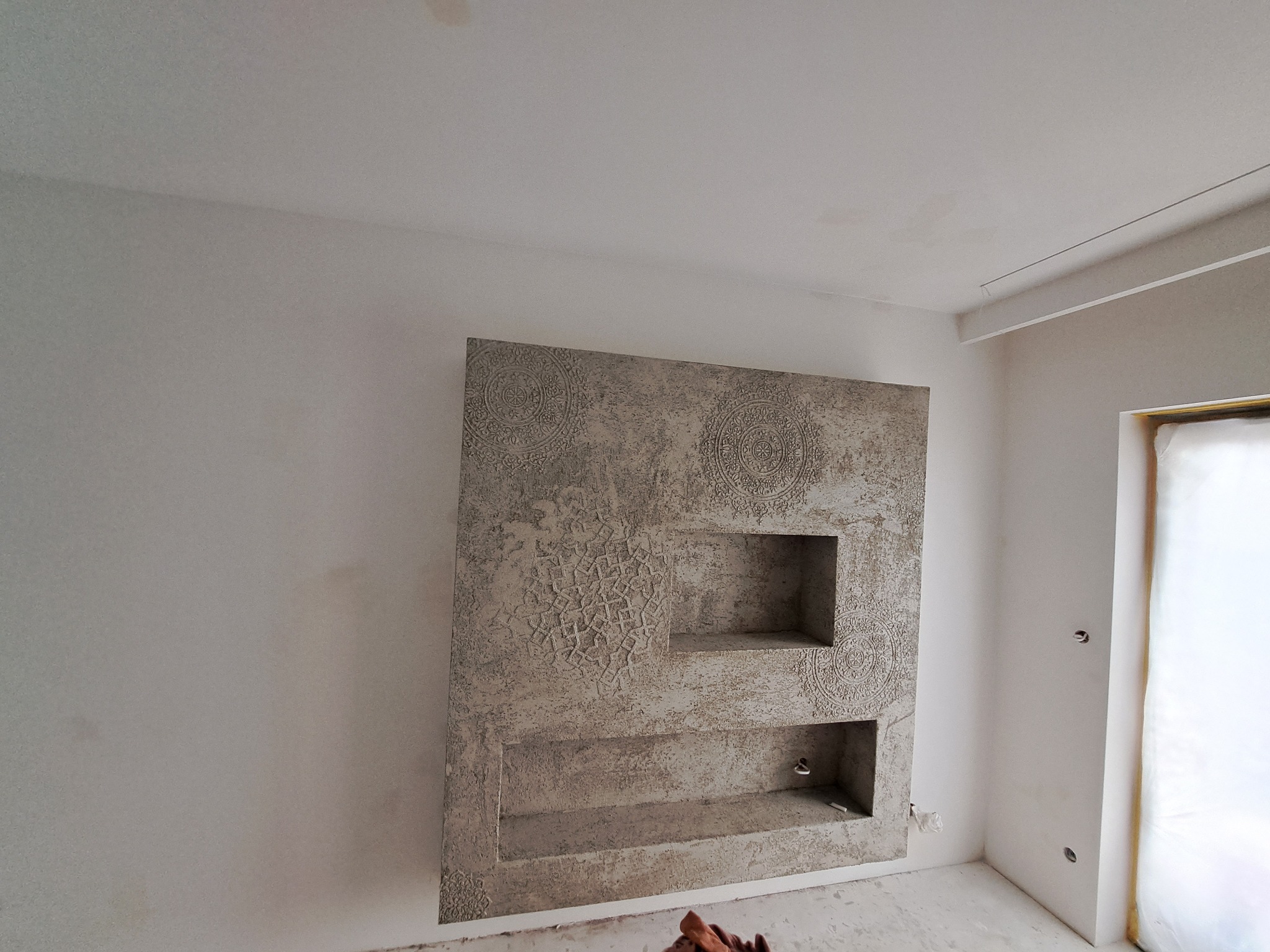 Ściana z dekoracyjną szpachlą o fakturze z wnękami, wzory geometryczne i motywy mandali, widoczne ślady prac remontowych.