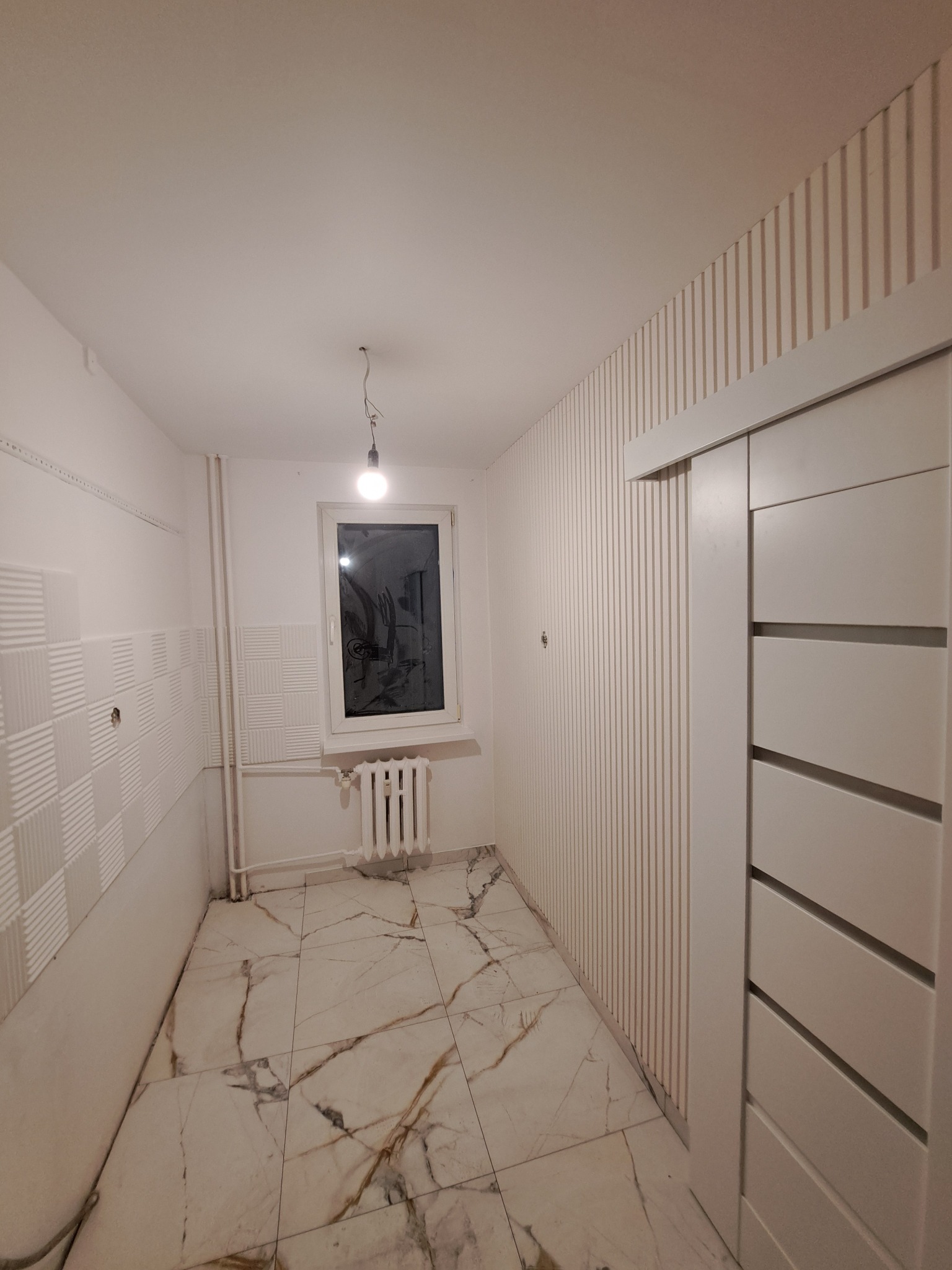 Wnętrze pokoju po remoncie: białe ściany z dekoracyjnymi panelami pionowymi i kwadratowymi, marmurowa podłoga, białe drzwi przesuwne i okno z grzejnikiem.