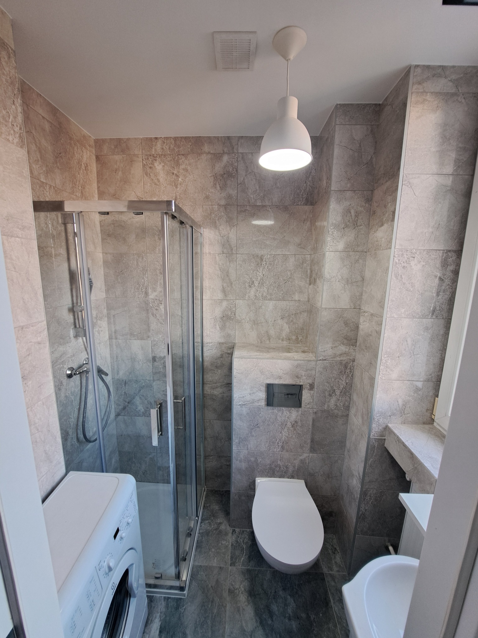 Wnętrze nowoczesnej łazienki z kabiną prysznicową, pralką, umywalką i podwieszaną toaletą, wykończonej szarymi płytkami imitującymi marmur, oświetlonej pojedynczą lampą wiszącą.