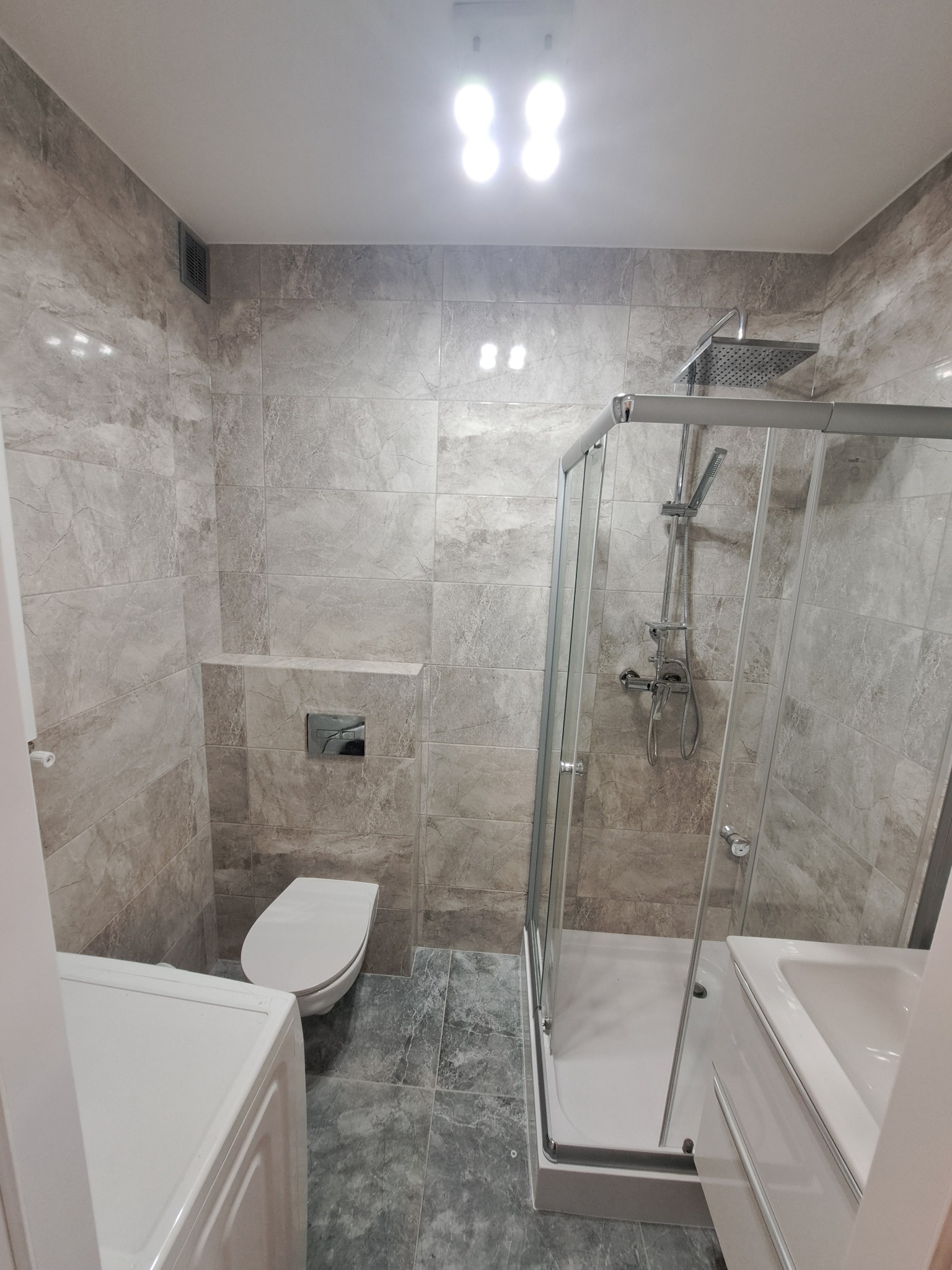 Nowoczesna łazienka po remoncie z kabiną prysznicową, podwieszaną toaletą i płytkami imitującymi marmur, oświetlona górnym światłem.