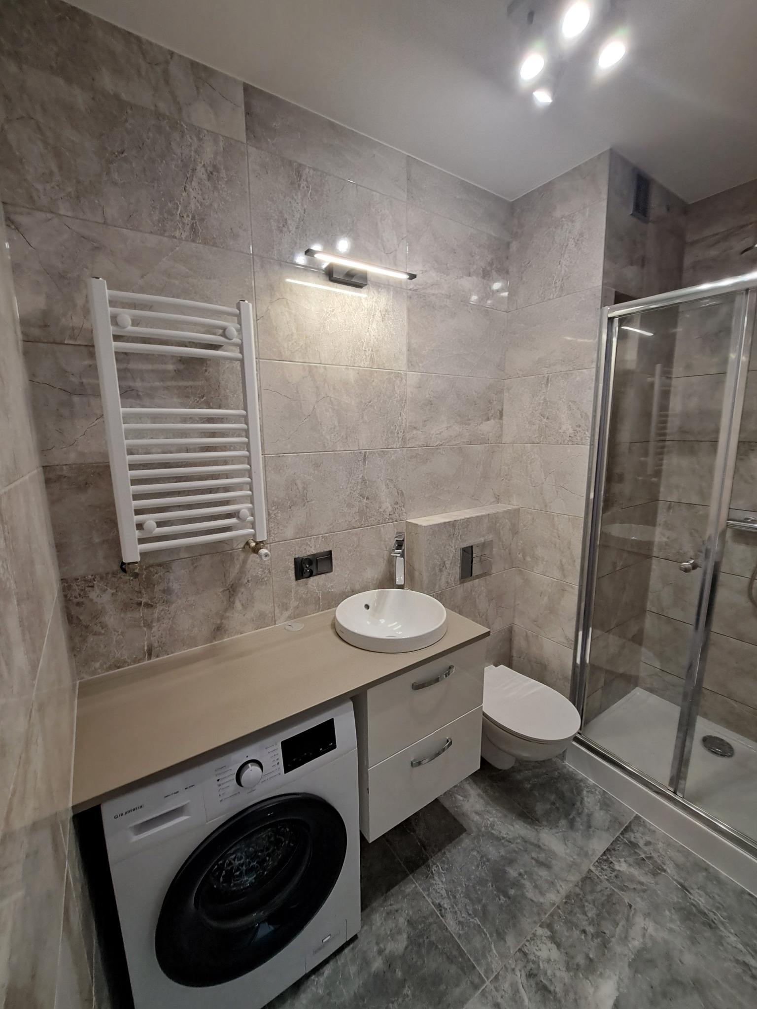 Nowoczesna łazienka w szarościach z pralką pod blatem, umywalką nablatową, grzejnikiem drabinkowym i kabiną prysznicową.