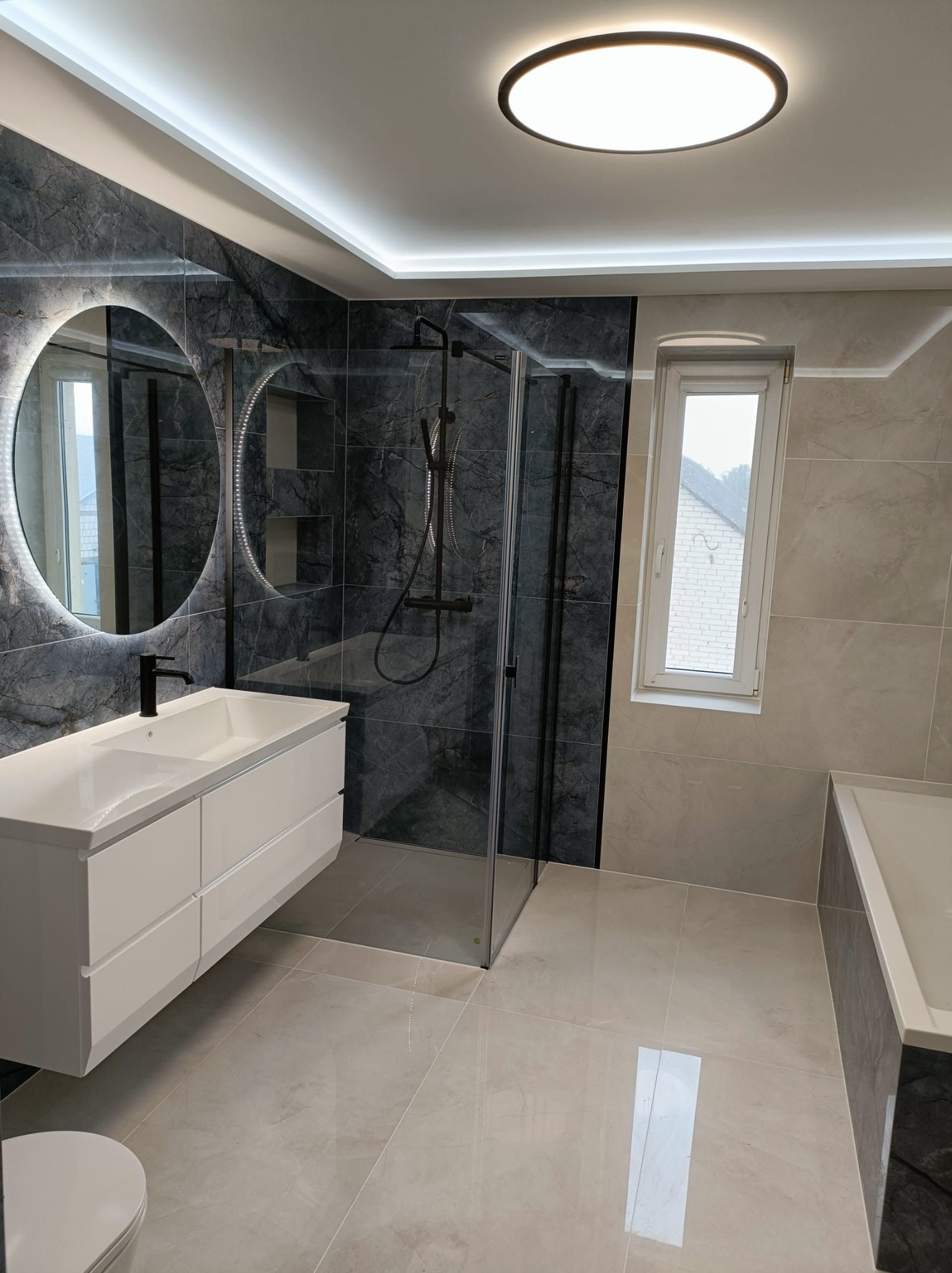Nowoczesna łazienka z szarymi płytkami imitującymi marmur, okrągłym lustrem z podświetleniem LED, białą szafką pod umywalką z czarną armaturą i przeszkloną kabiną prysznicową.