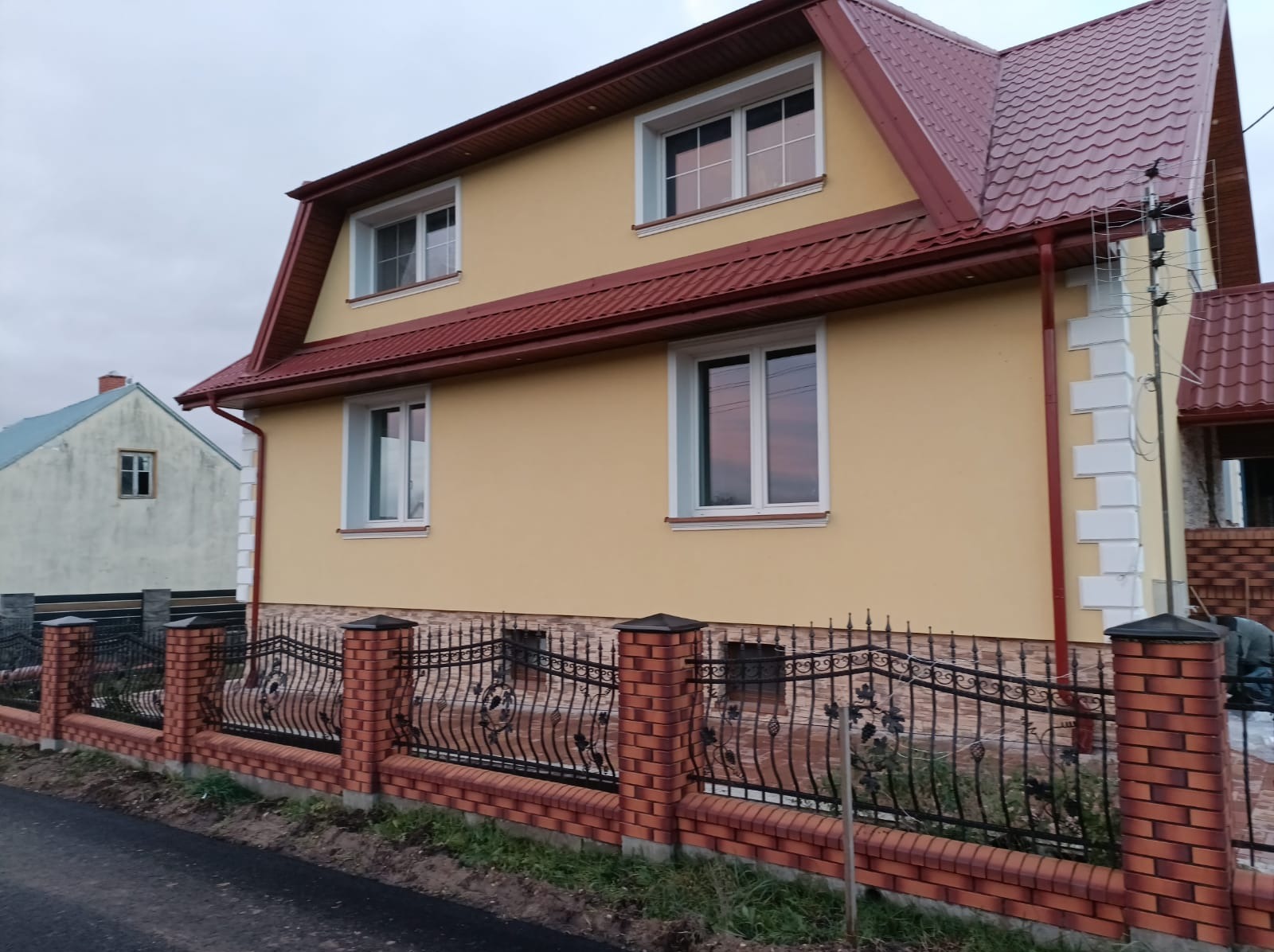 Żółty dom z czerwoną dachówką i ozdobnym, kutym ogrodzeniem na tle pochmurnego nieba.