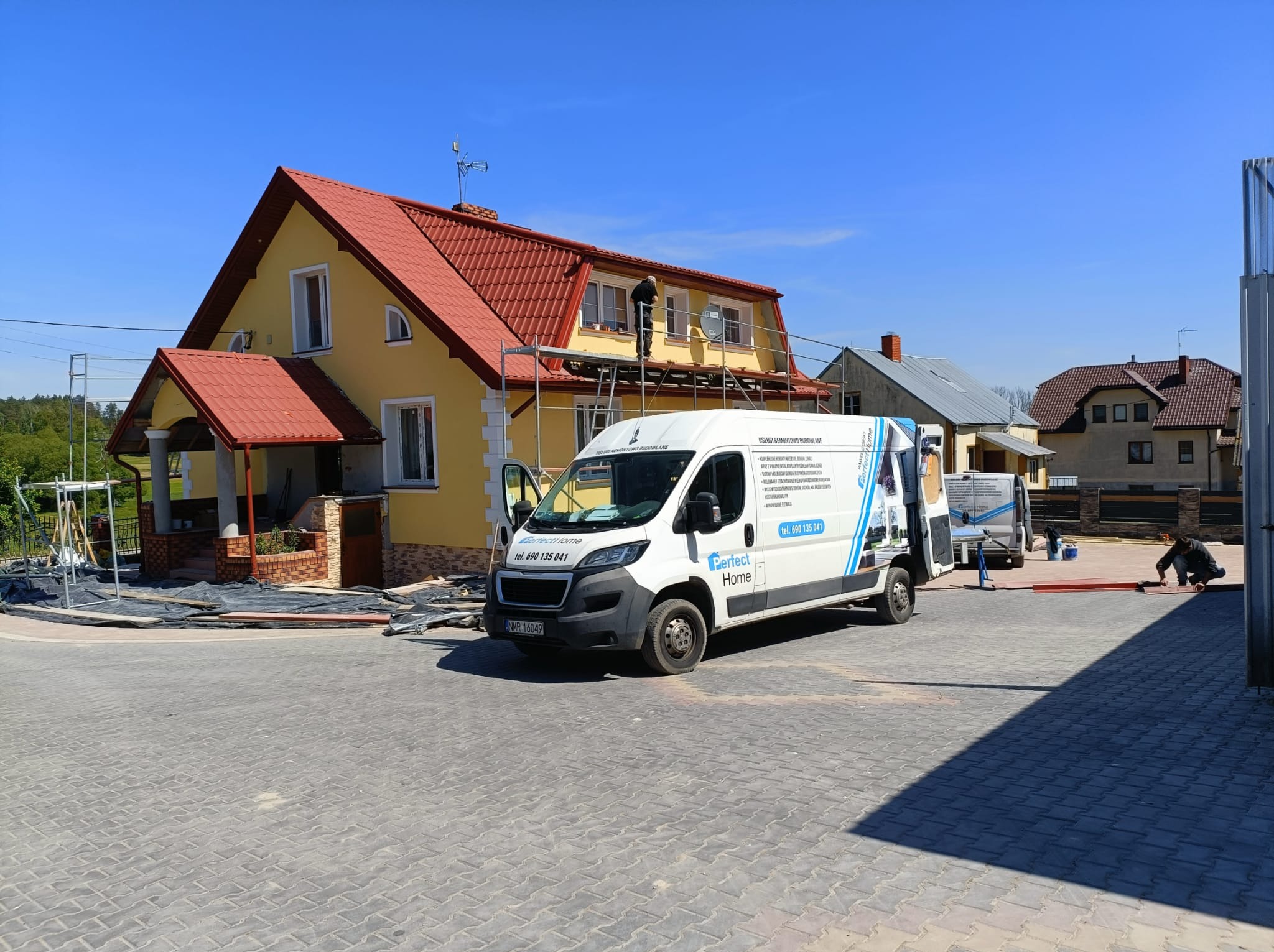 Żółty dom jednorodzinny z czerwoną dachówką, ocieplany z zewnątrz, pracownik na rusztowaniu, firmowy bus Perfect Home na kostce brukowej, błękitne niebo.