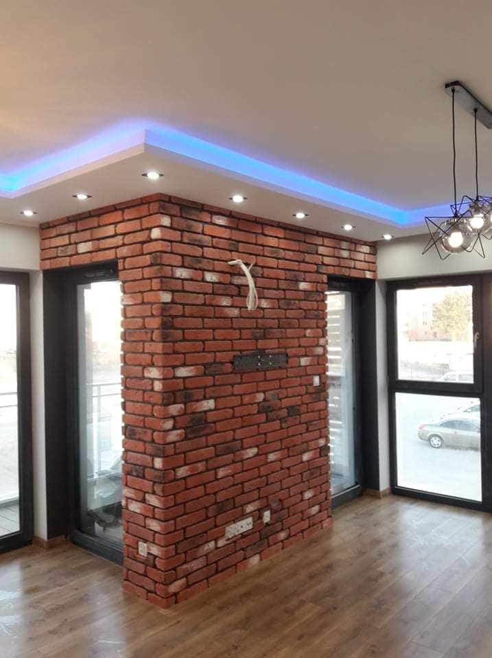 Fragment ściany wykończony czerwoną cegłą klinkierową w nowoczesnym wnętrzu z parkietem, oświetleniem LED i czarnymi ramami okiennymi.