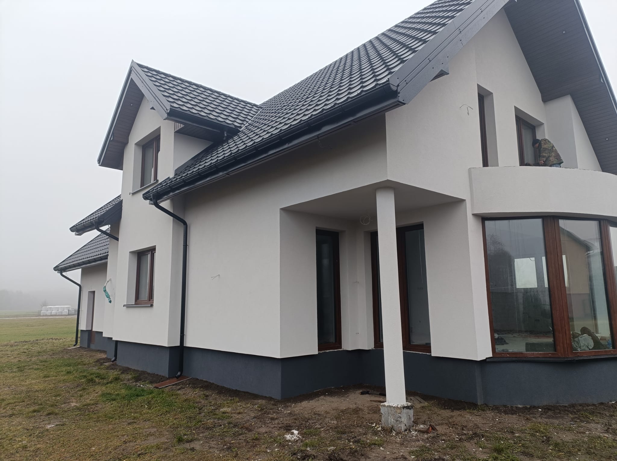 Nowoczesny dom jednorodzinny z białymi ścianami i grafitowym dachem, widoczny z zewnątrz w pochmurny dzień, z pracownikiem na balkonie.