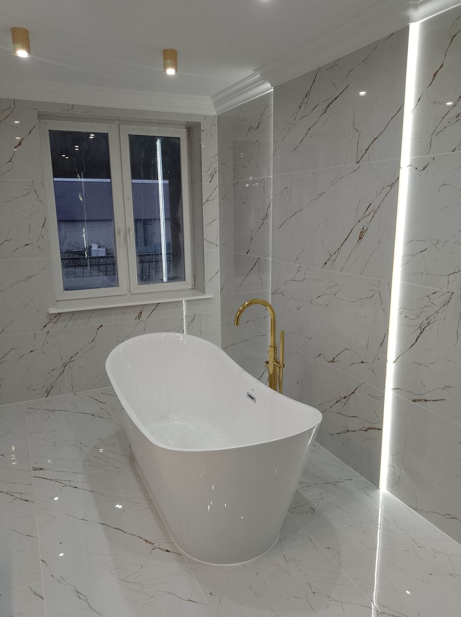 Nowoczesna łazienka z wolnostojącą białą wanną, złotą armaturą i marmurowymi płytkami z brązowymi żyłami oraz oświetleniem LED.
