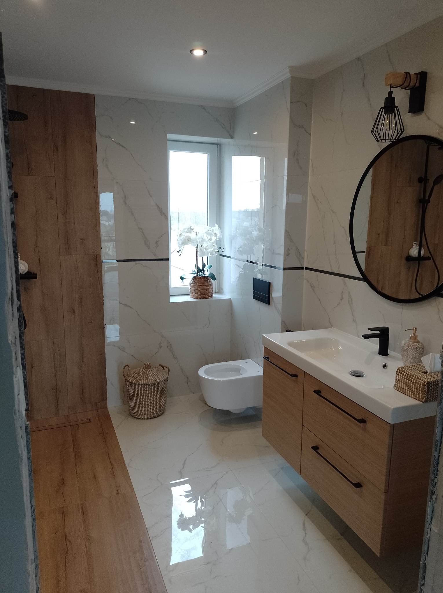 Nowoczesna łazienka z drewnianymi akcentami: panele prysznicowe imitujące drewno, marmurowe płytki, okrągłe lustro z czarną ramą i szafka pod umywalkę z drewnianymi frontami i czarnymi uchwytami.