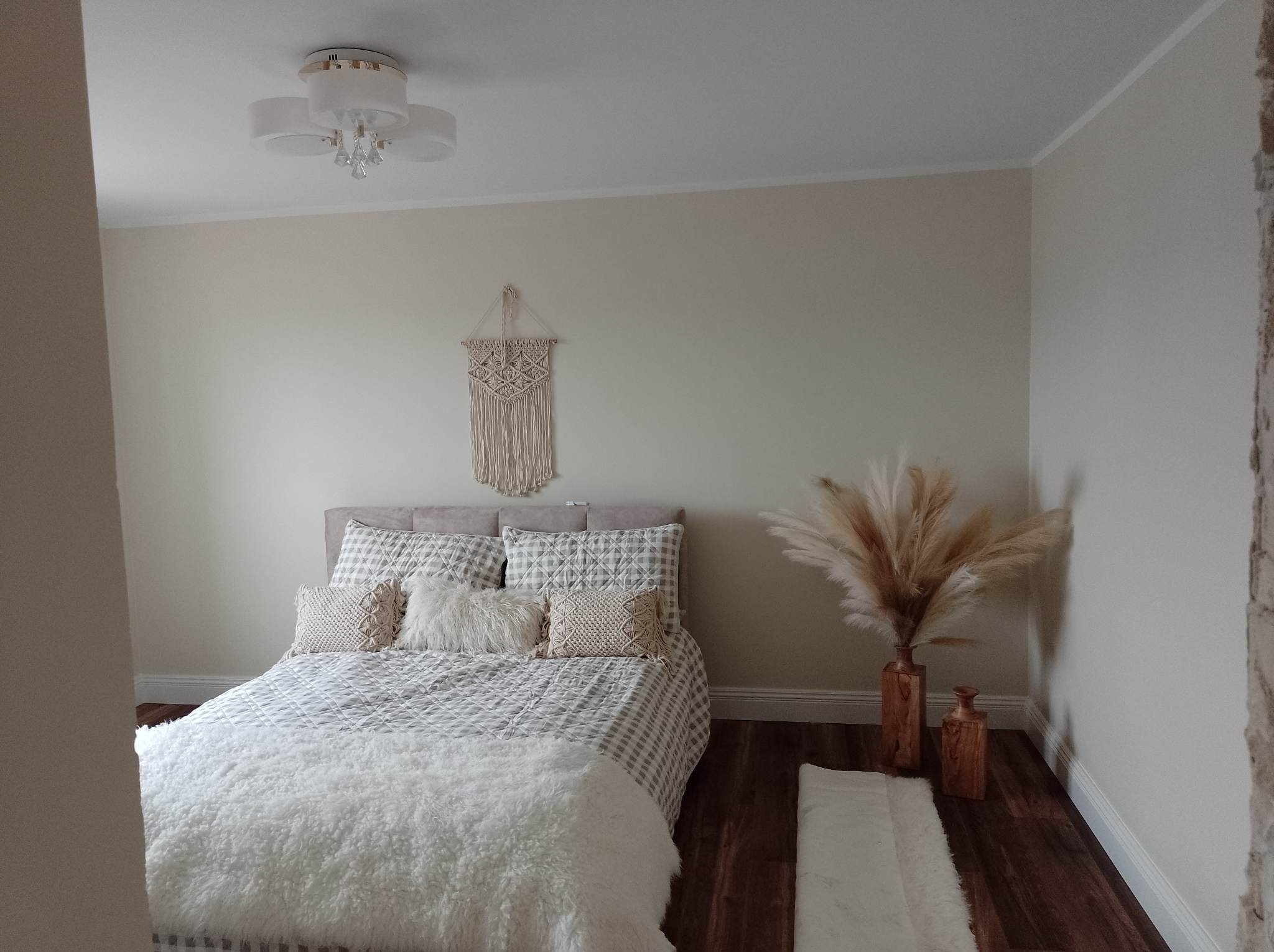 Jasna sypialnia z łóżkiem w kratę, puszystym pledem i dekoracjami z trawy pampasowej w drewnianych wazonach, na ścianie makrama w stylu boho.