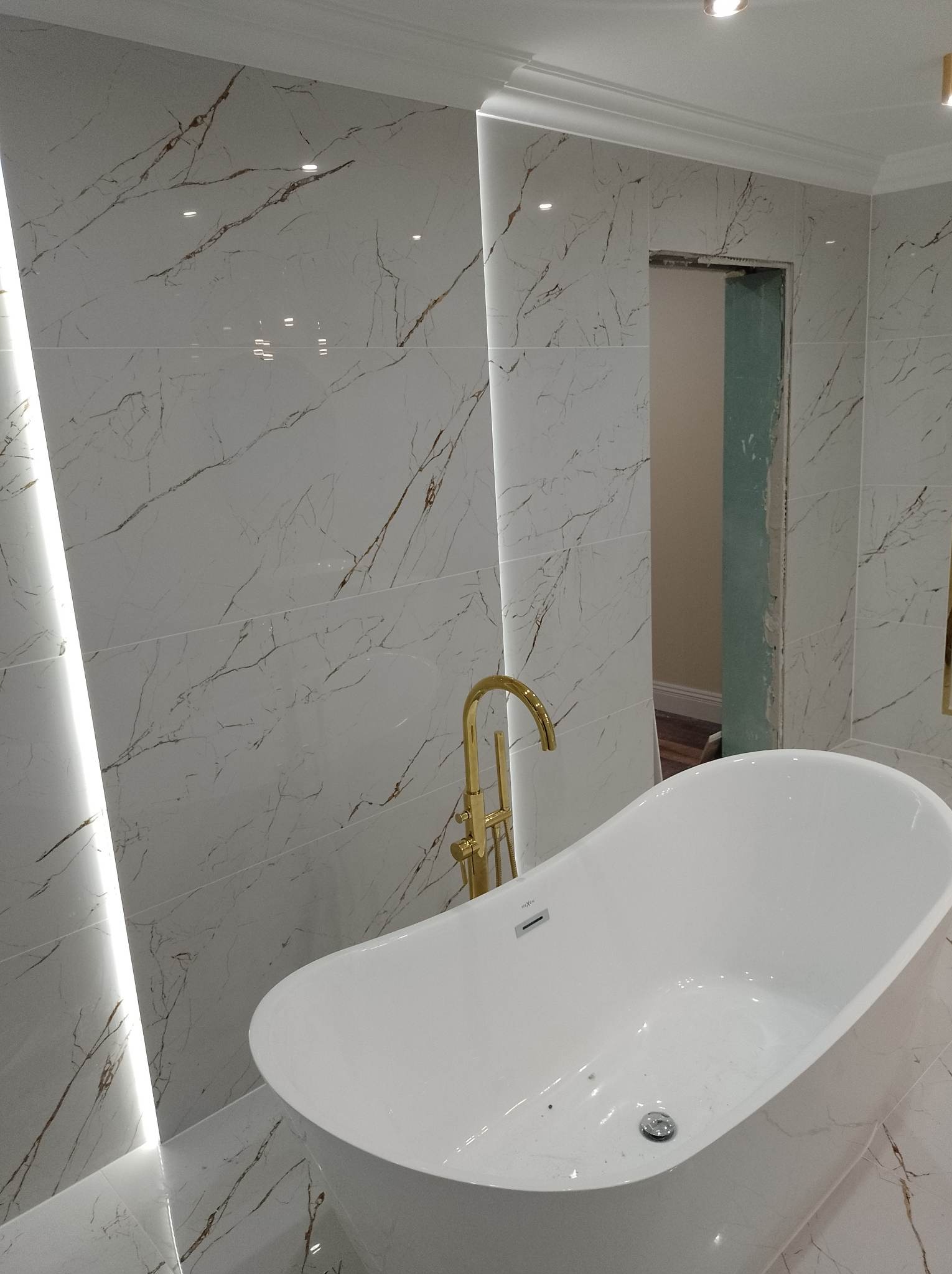 Nowoczesna łazienka w Wiznie z wolnostojącą białą wanną i złotą armaturą na tle ściany wyłożonej białymi płytkami imitującymi marmur z podświetleniem LED.