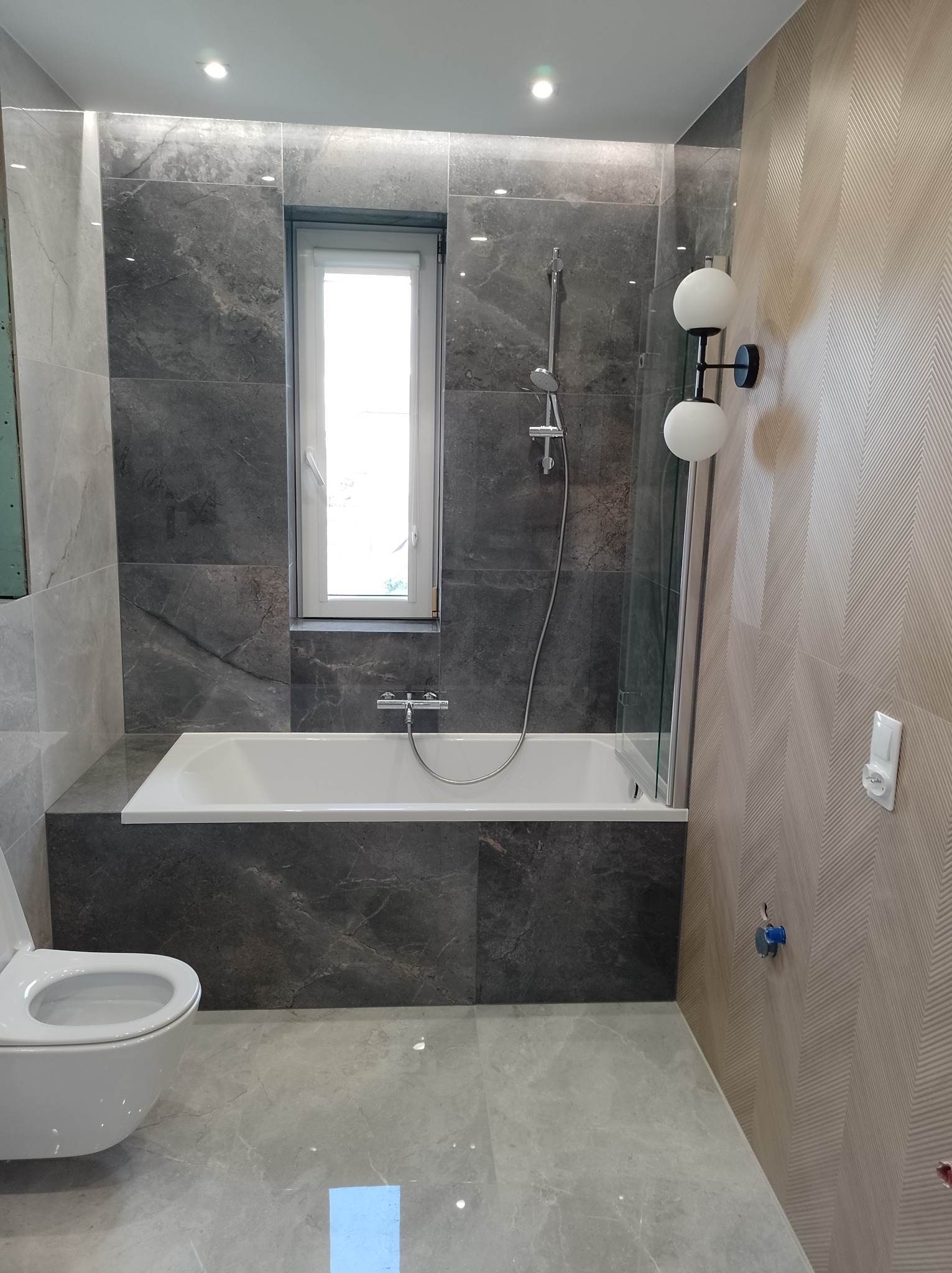 Nowoczesna łazienka z szarymi marmurowymi płytkami, białą wanną i toaletą, oraz lampą ścienną z kulistymi kloszami. Okno nad wanną wpuszcza naturalne światło.