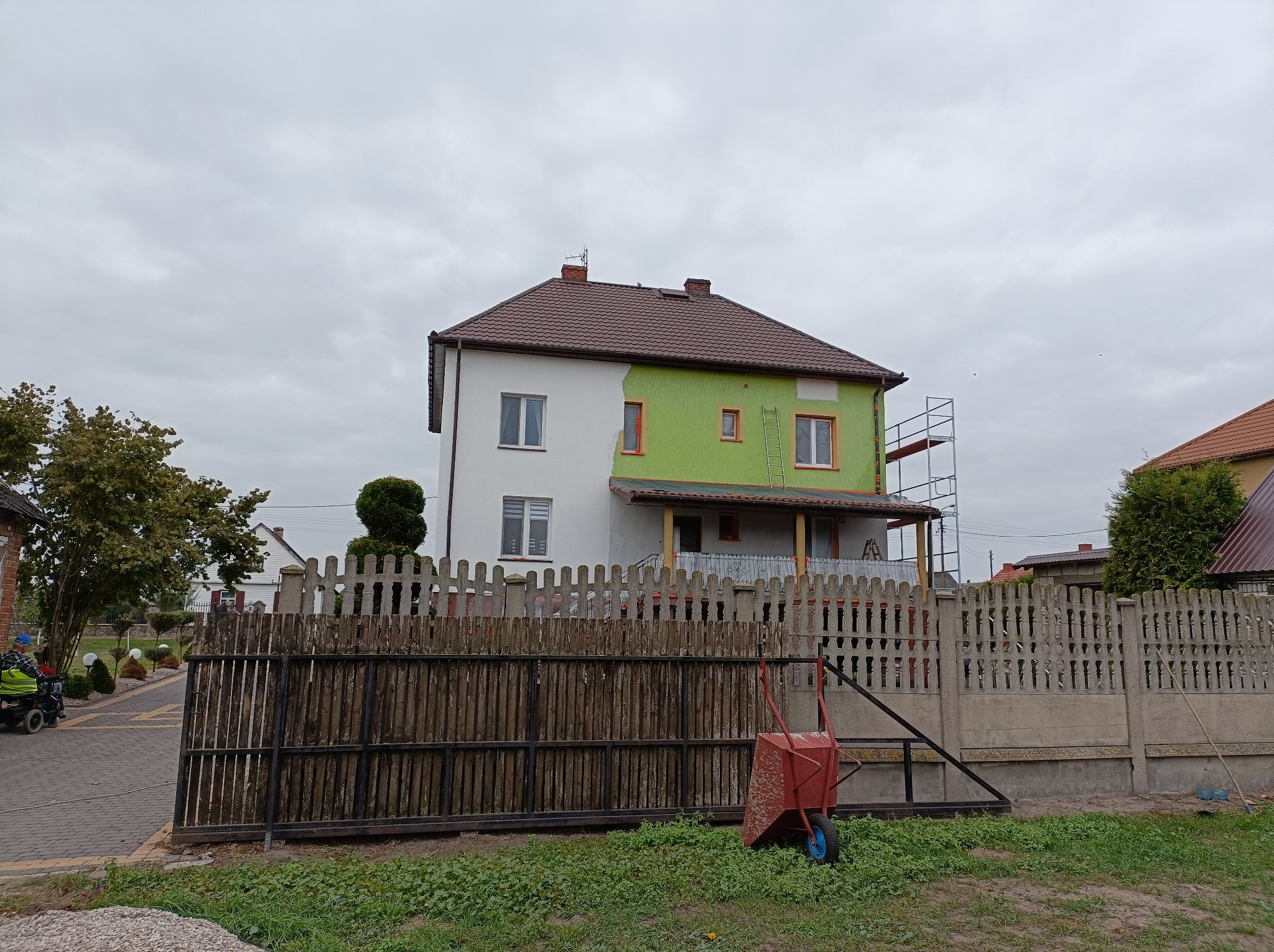 Dwukondygnacyjny dom z brązowym dachem, częściowo pomalowany na biało, a częściowo na zielono, z rusztowaniem i drabiną przy elewacji, widoczny zza drewnianego płotu i czerwonej taczki.