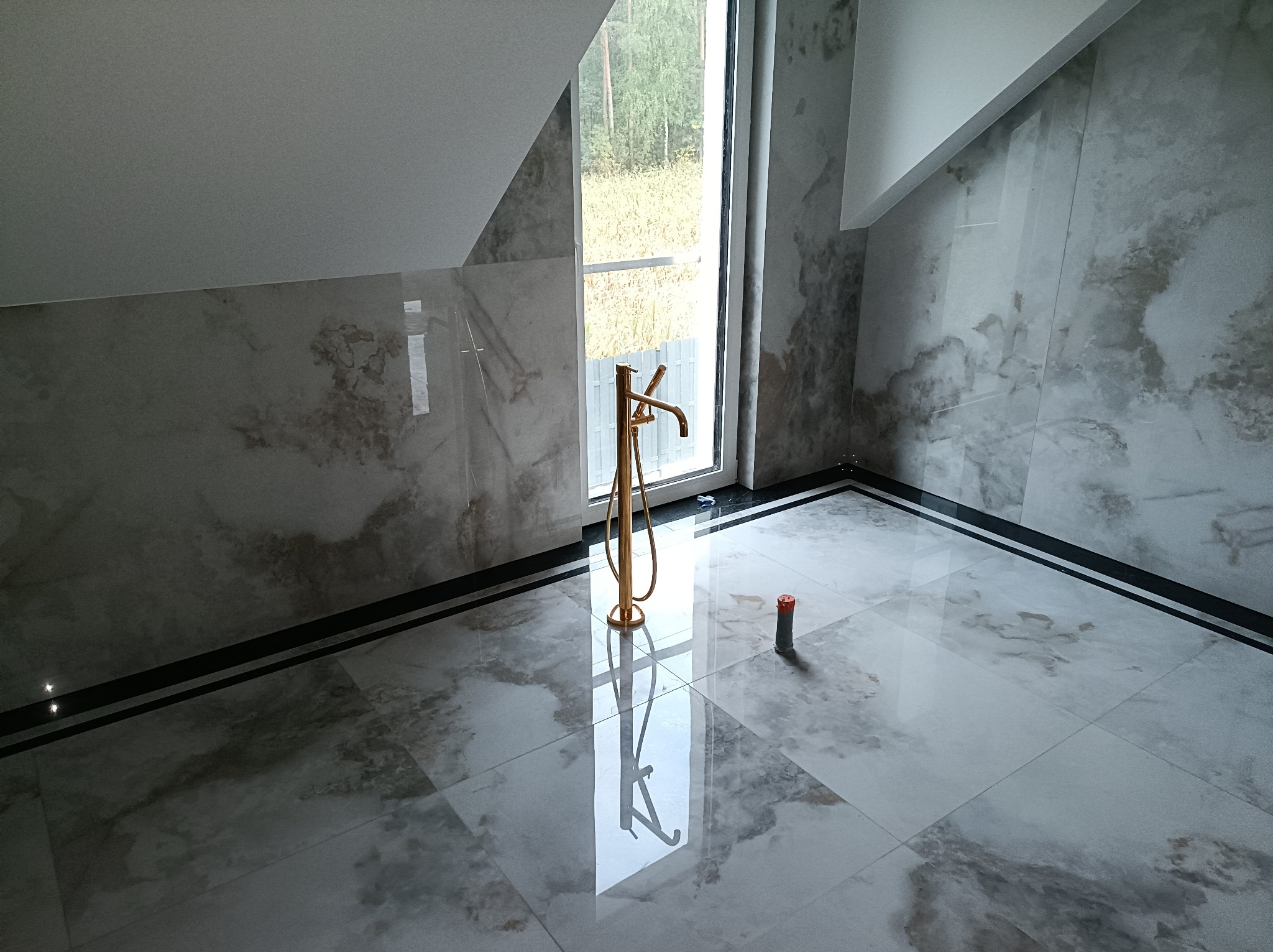 Nowoczesna łazienka na poddaszu z marmurowymi płytkami, złotą baterią wolnostojącą i oknem z widokiem na zieleń.