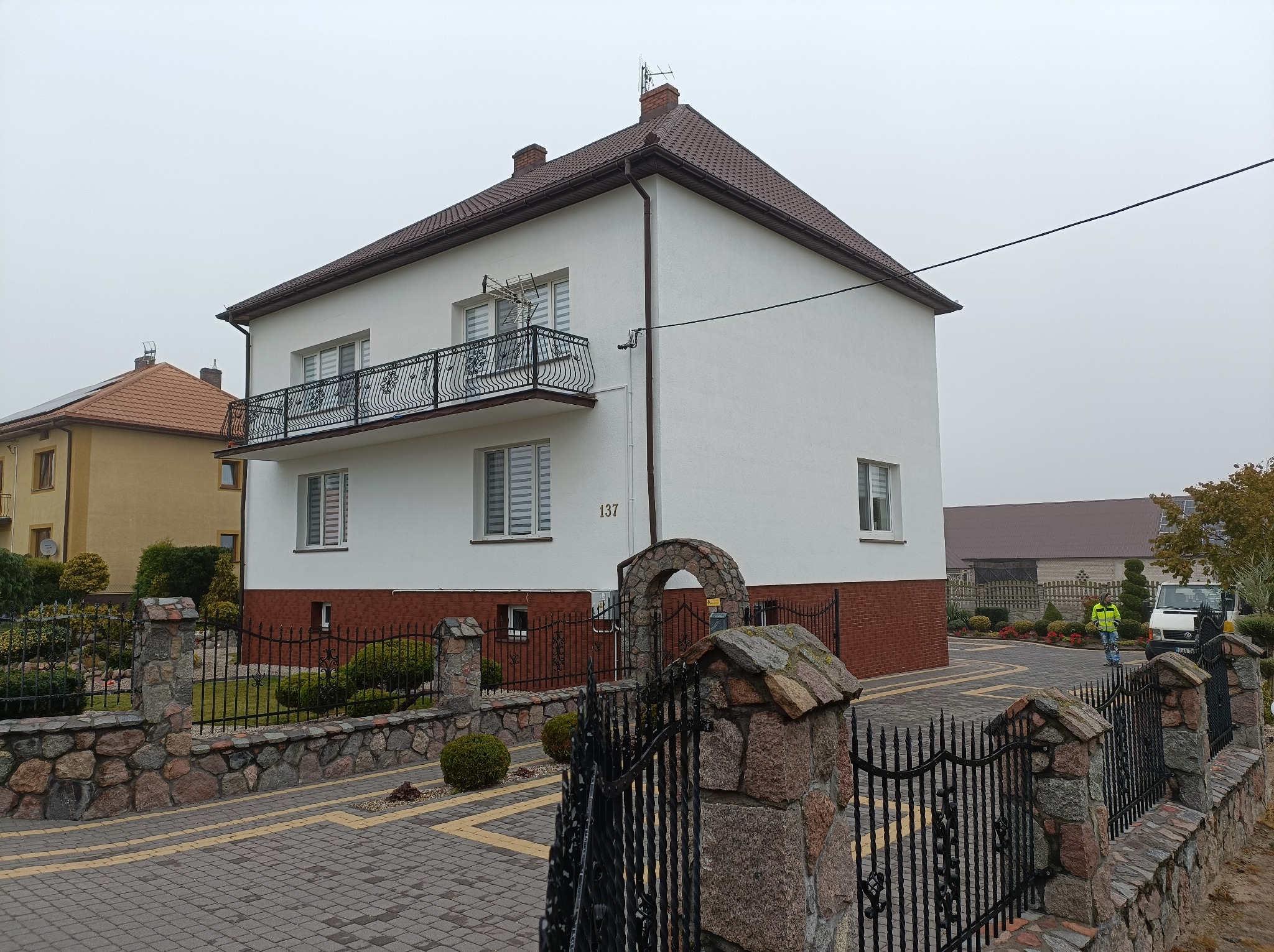 Dwupiętrowy dom z białymi ścianami i brązowym dachem, z balkonem z kutą balustradą, widoczny zza kamienno-metalowego ogrodzenia z bramą.