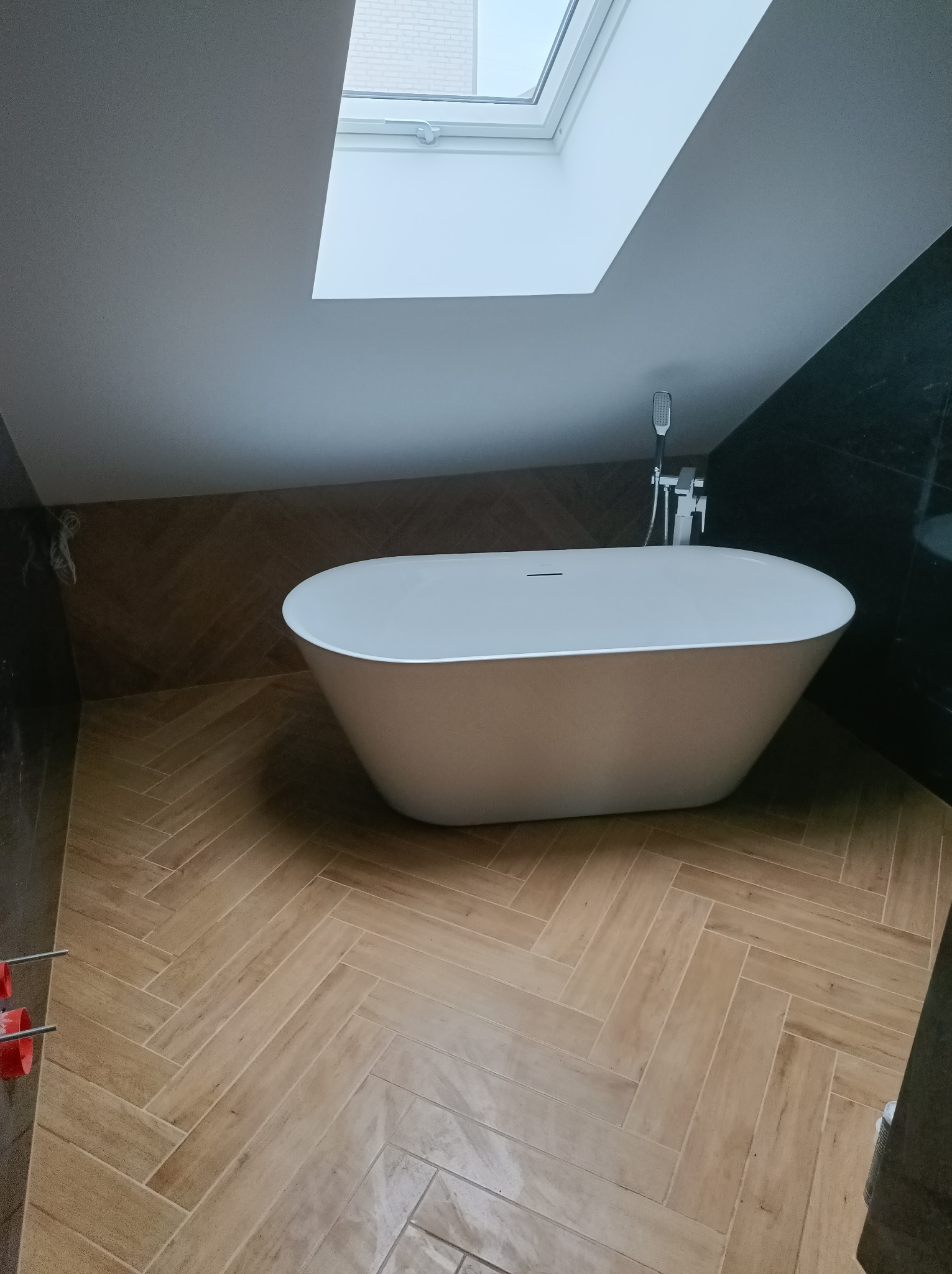 Wolnostojąca wanna owalna w nowoczesnej łazience na poddaszu z oknem dachowym, wykończona płytkami imitującymi drewno ułożonymi w jodełkę.