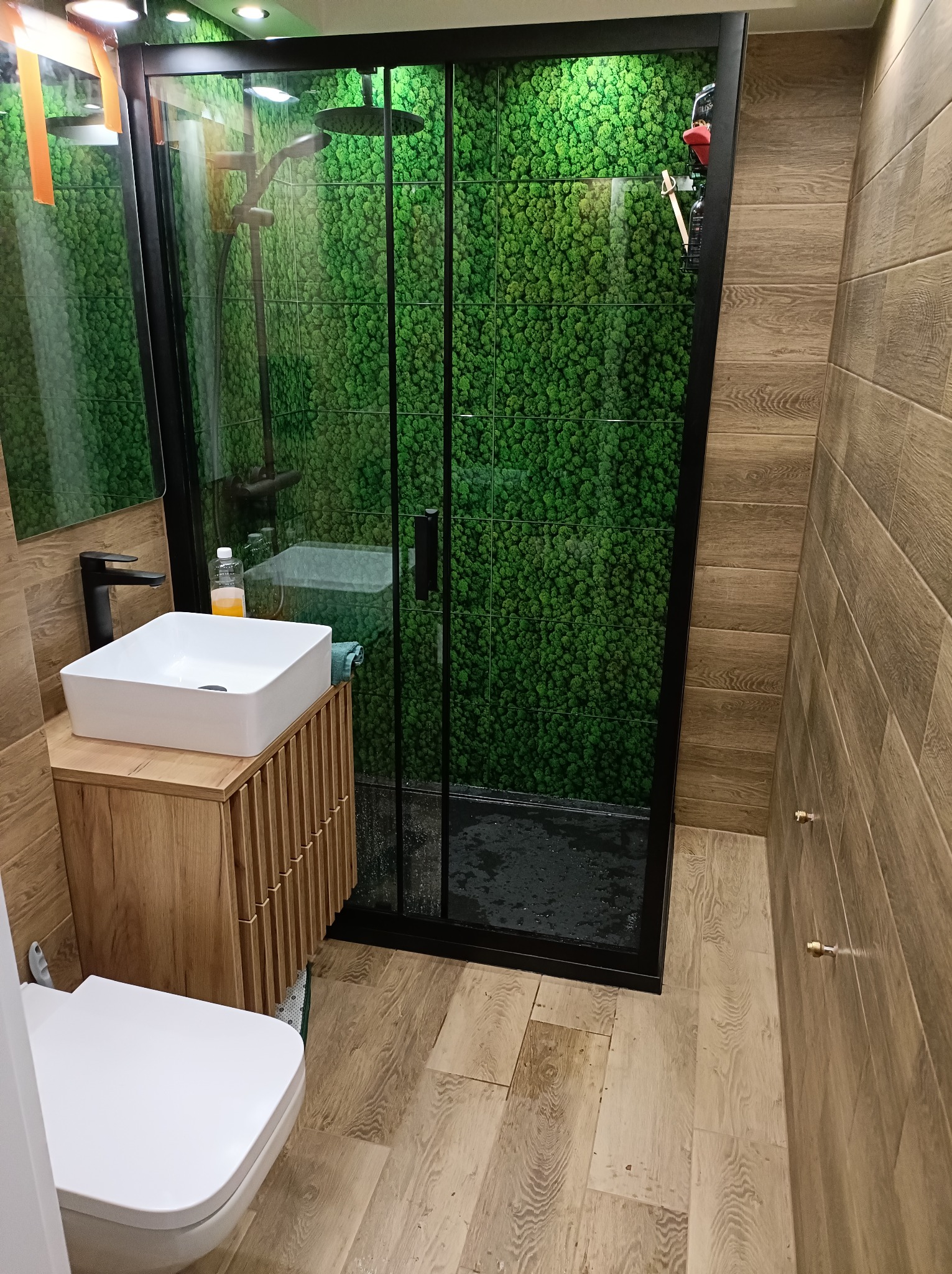 Nowoczesna łazienka z kabiną prysznicową z czarną ramą i ścianą z mchu, drewnopodobne płytki na ścianach i podłodze, biała umywalka nablatowa na drewnianej szafce z lamelami, wisząca toaleta.