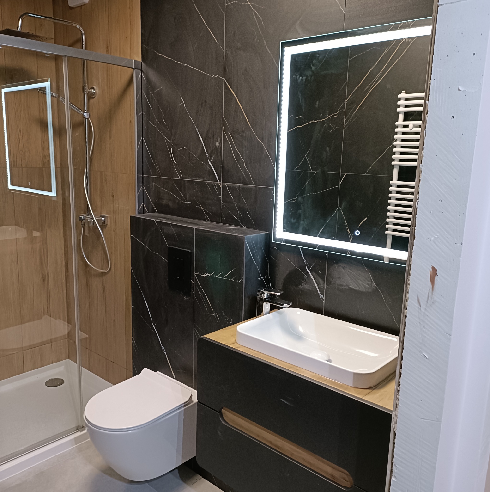 Nowoczesna łazienka z kabiną prysznicową, podwieszaną toaletą i umywalką nablatową na szafce z drewnianymi akcentami, oświetlone lustro na tle czarnych płytek imitujących marmur, widoczny fragment...