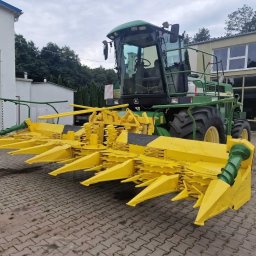 Wiktor Kawa - Zielono-żółty kombajn rolniczy John Deere stoi na brukowanym placu przed budynkiem, prezentując zamontowany heder do zbioru kukurydzy z charakterystycznymi, żółtymi stożkami.