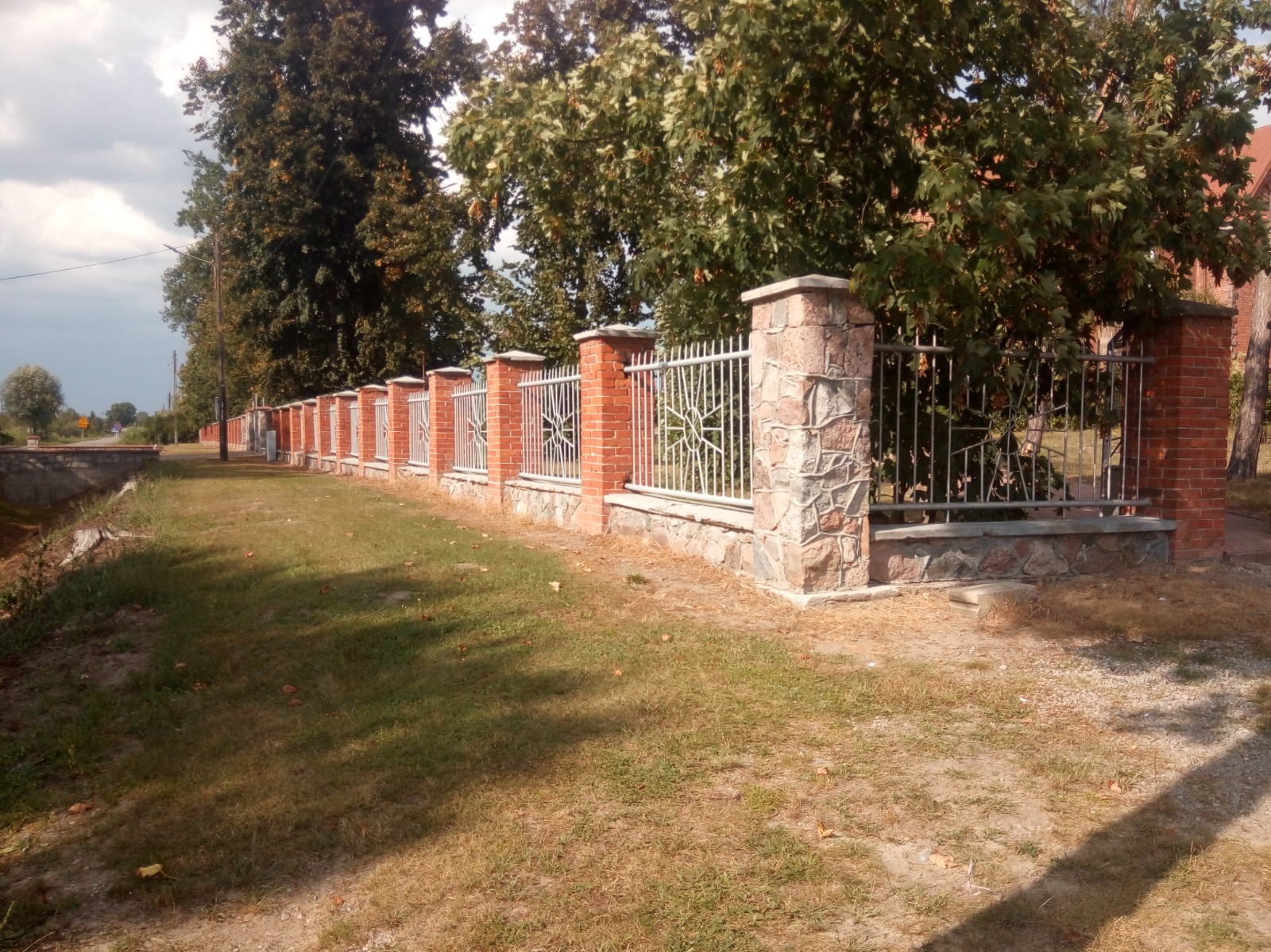 Długa linia ogrodzenia z cegły i kamienia z metalowymi przęsłami o dekoracyjnym wzorze, ciągnąca się wzdłuż drogi, z drzewami w tle.