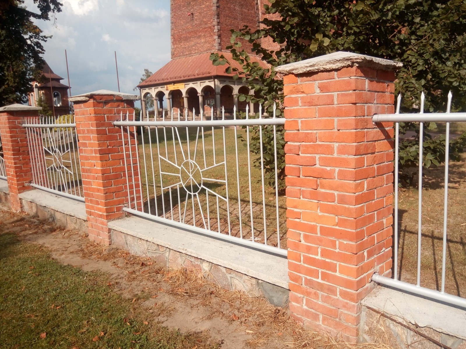 Fragment ogrodzenia z ceglanymi słupkami i metalowymi przęsłami z dekoracyjnym motywem koła z promieniami, na tle trawnika i budynków.