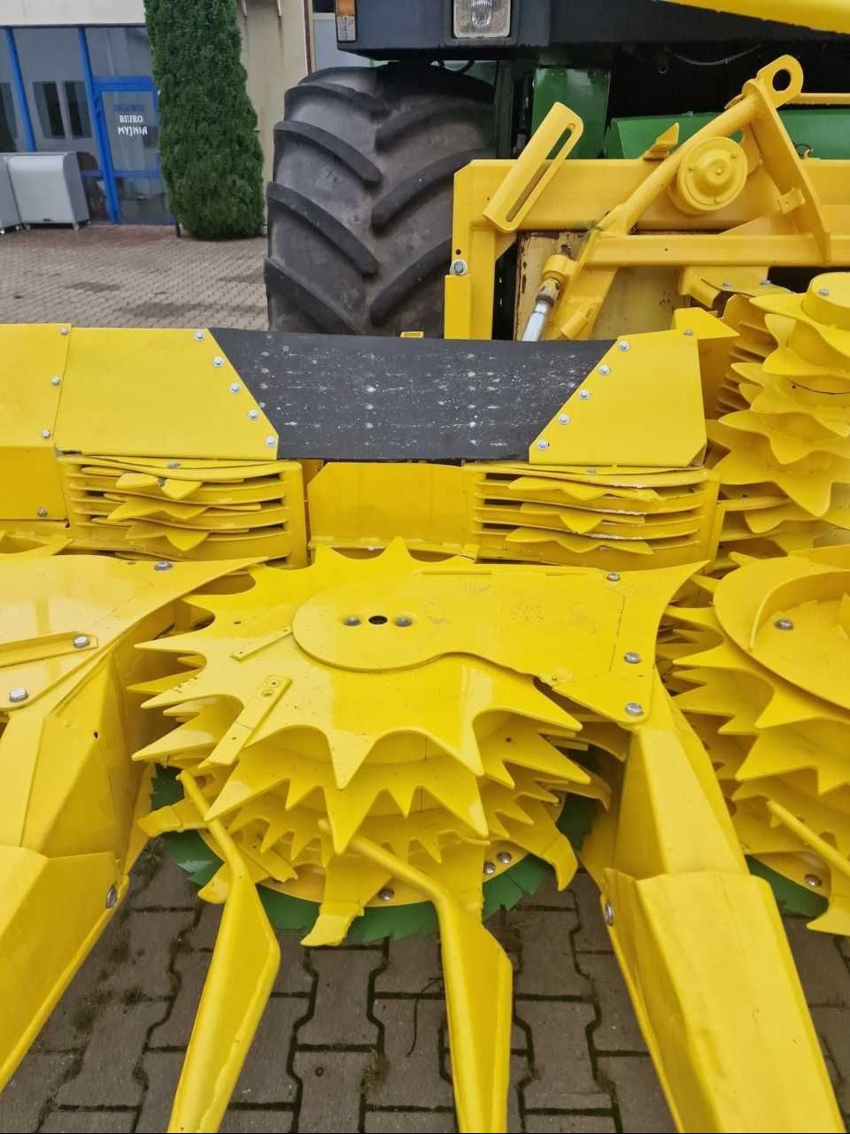 Żółty, metalowy sprzęt rolniczy z zębatymi elementami tnącymi, zamontowany z tyłu traktora, widoczny na brukowanym placu.
