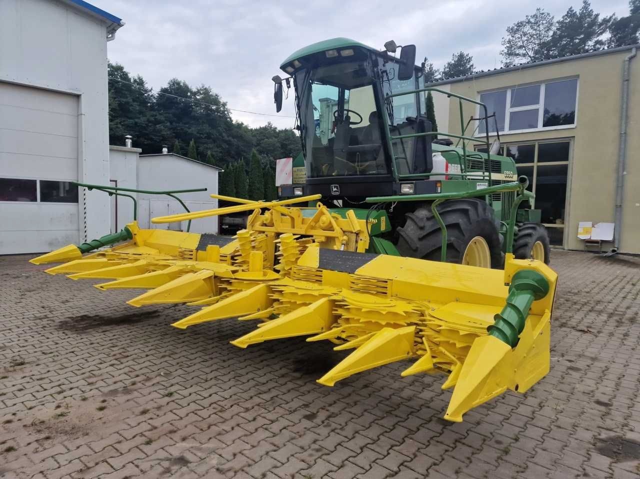Zielono-żółty kombajn rolniczy John Deere stoi na brukowanym placu przed budynkiem, prezentując zamontowany heder do zbioru kukurydzy z charakterystycznymi, żółtymi stożkami.
