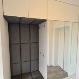 Biała zabudowa przedpokoju z siedziskiem obitym szarą tkaniną w kwadraty, wbudowane lustro oraz p&oacute;łka na buty.