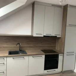 Nowoczesna kuchnia w Krakowie z białymi szafkami i drewnianym blatem, dopasowana do skosu dachu, z czarną armaturą i płytą indukcyjną.