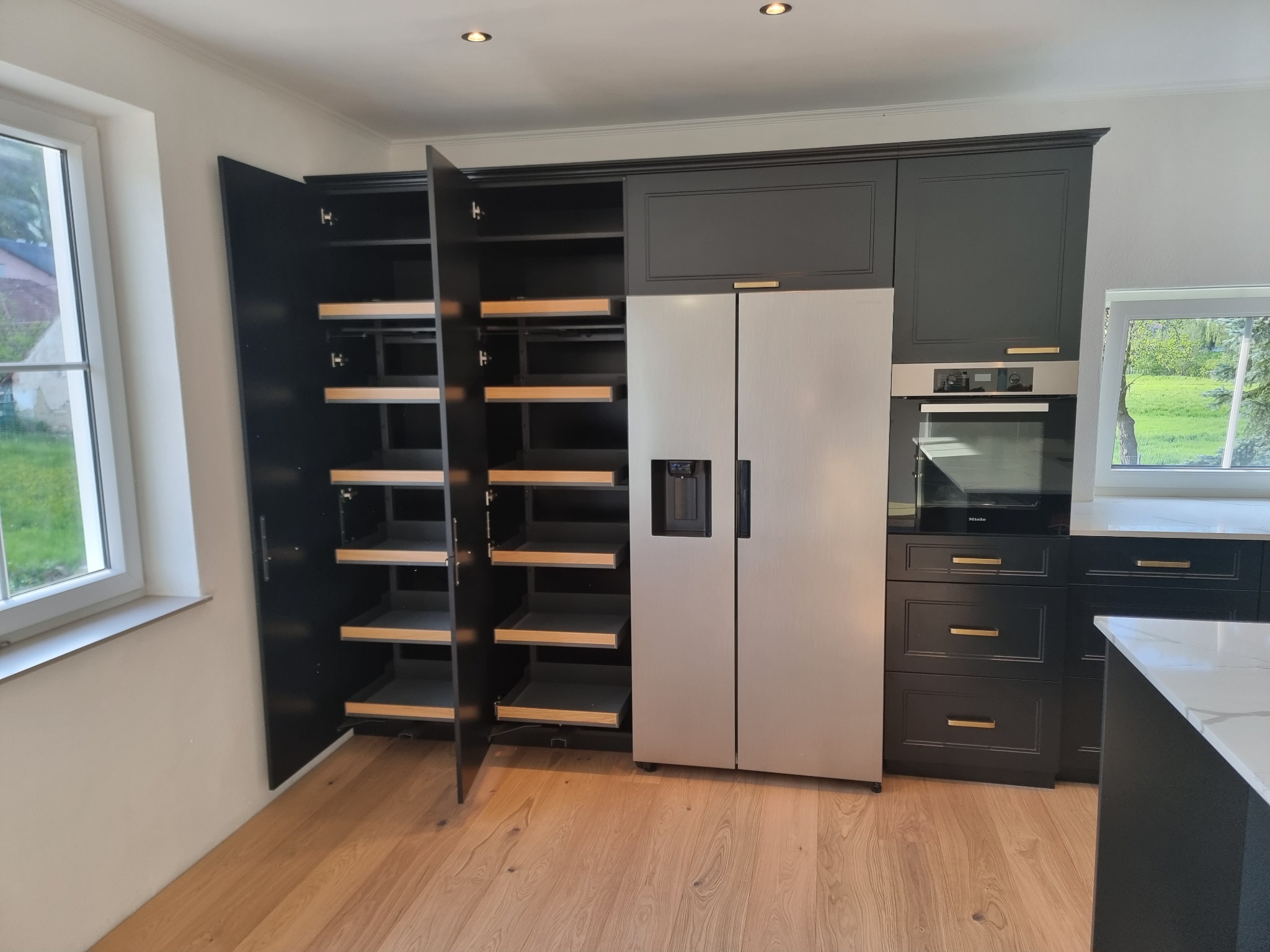 Czarna zabudowa kuchenna z szufladami cargo w otwartej szafce obok srebrnej lodówki side-by-side, widok na jasną podłogę i okno.